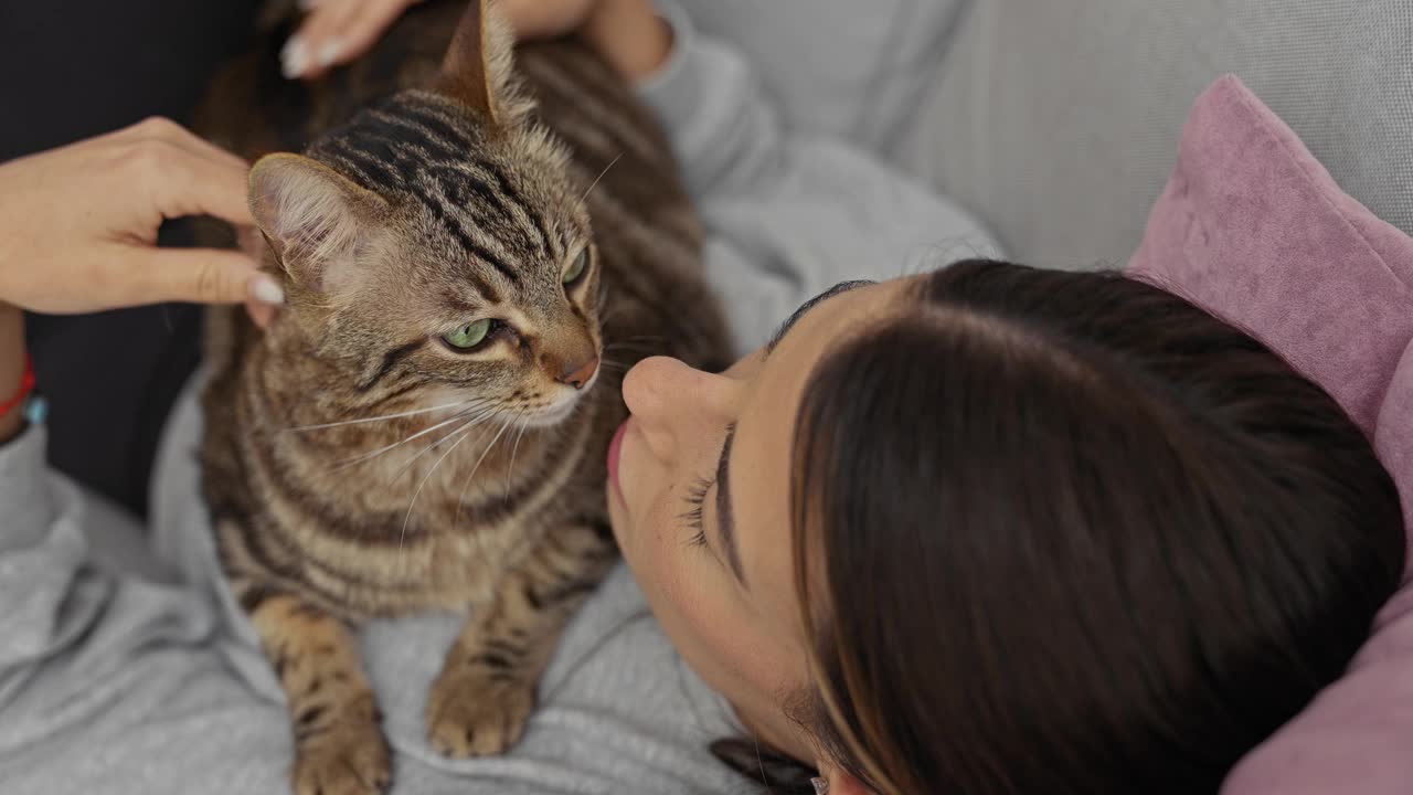 一位年轻女子躺在客厅里，深情地抚摸着坐在她胸前的虎斑猫，营造出一种舒适而亲密的家庭氛围。视频素材