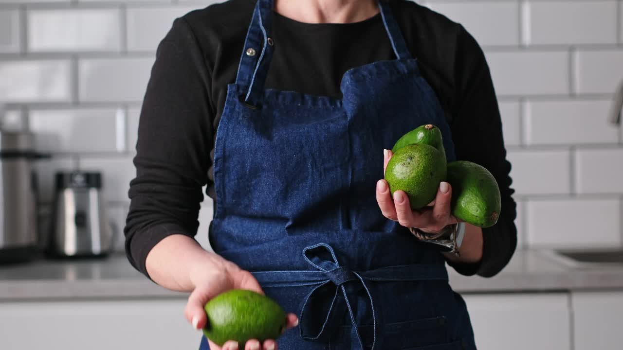 穿着围裙的女人在厨房里扔和抓新鲜的绿色鳄梨视频下载