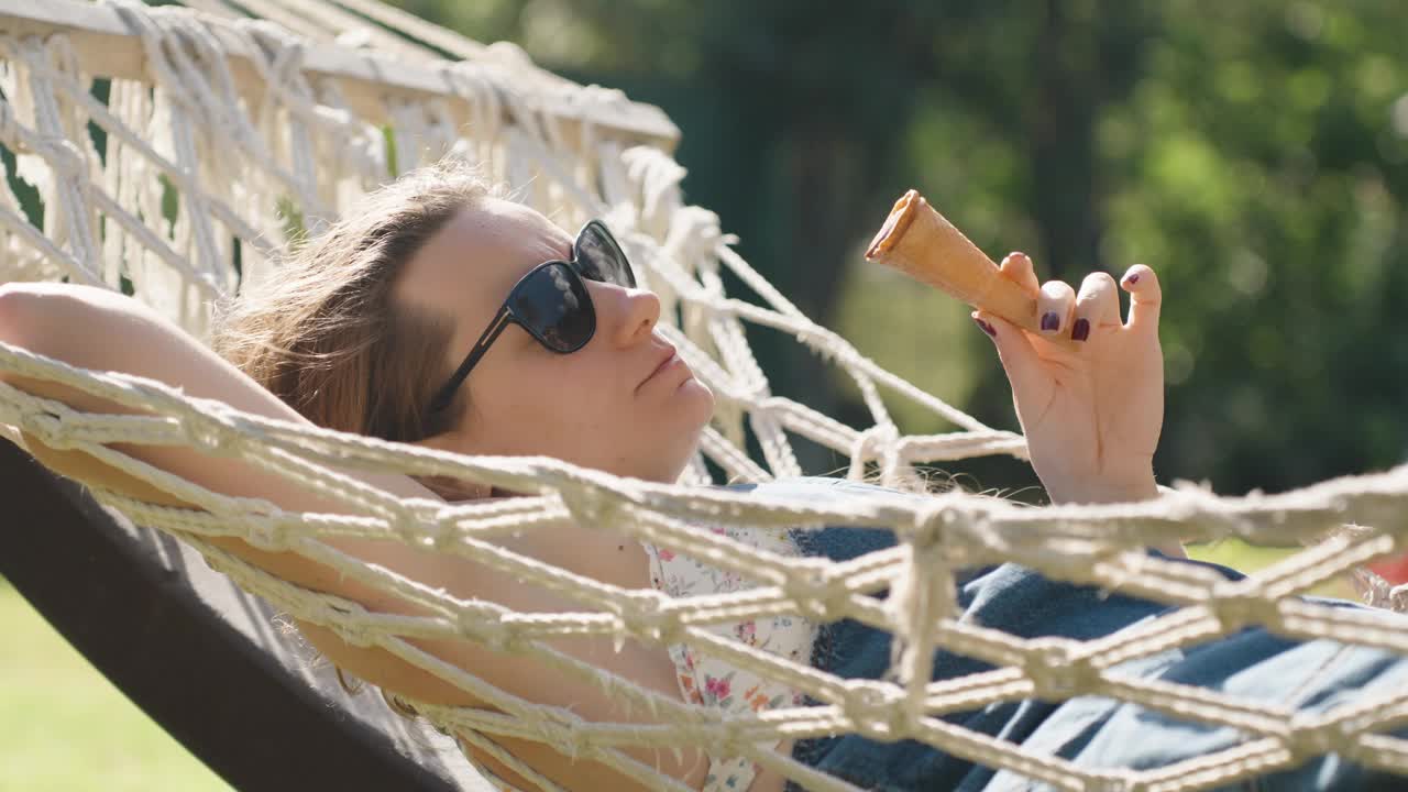 一位年轻女子躺在吊床上，一边晒太阳，一边吃着冰淇淋。视频素材