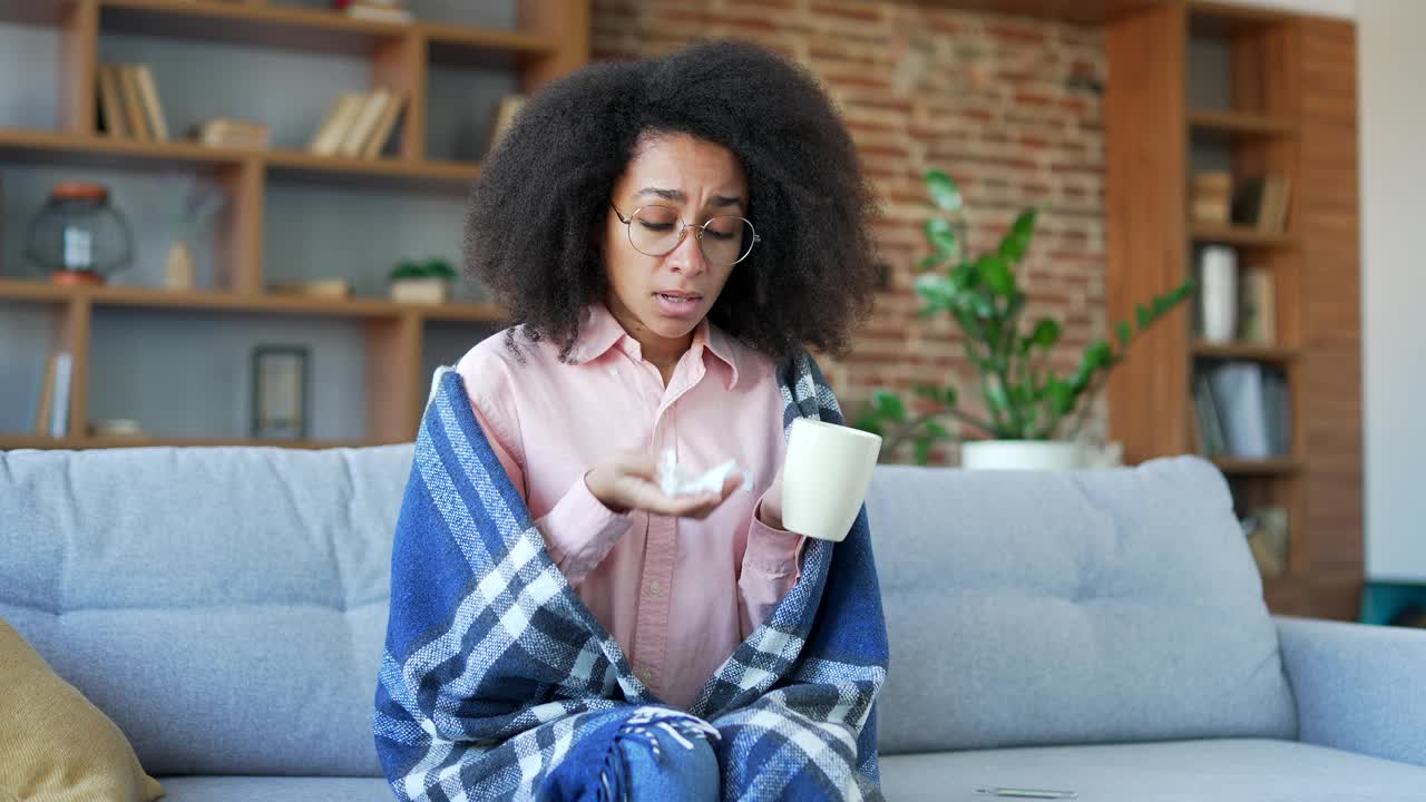 一位患有感冒和流感的非裔美国妇女坐在家里客厅的沙发上。视频素材