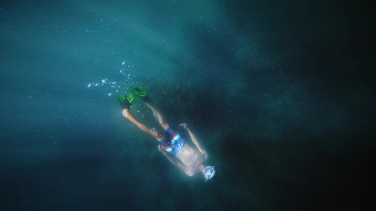 一只雄性潜水者在珊瑚礁附近的深海蓝色海水下游泳的俯视图。太阳的眩光和水下的直射阳光都是可见的视频素材