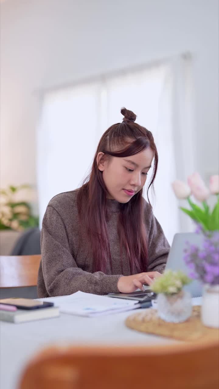 亚洲女商人在家工作，在客厅的桌子上使用她的笔记本电脑视频素材