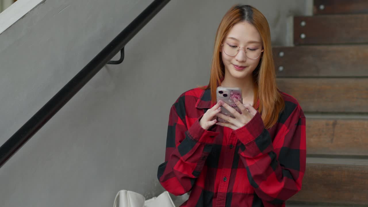 快乐的年轻Z世代女性坐在楼梯的台阶上，用她的手机与朋友和家人联系，网上购物，玩网络游戏，连接社交媒体视频素材