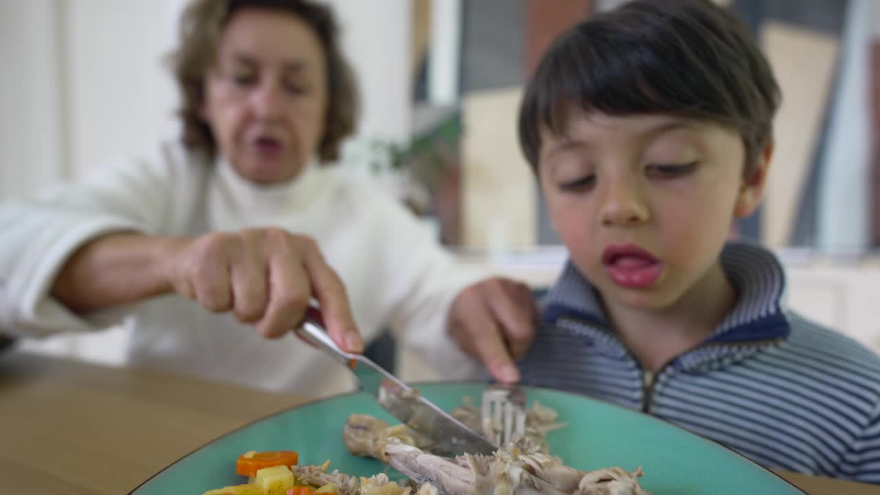 小男孩在祖母为他切好食物后，急切地享受着他的食物。男孩拿着叉子盛着食物，而祖母，有点模糊，还在餐桌旁视频素材