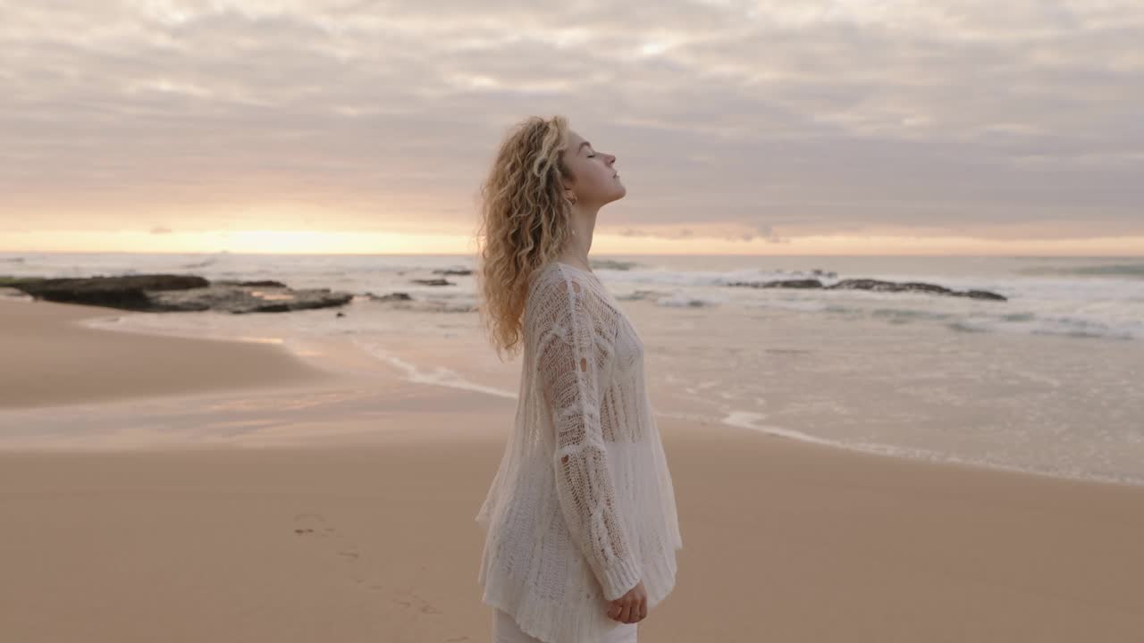 在日落的海滩上，穿着白色连衣裙的女人感受着海风视频素材
