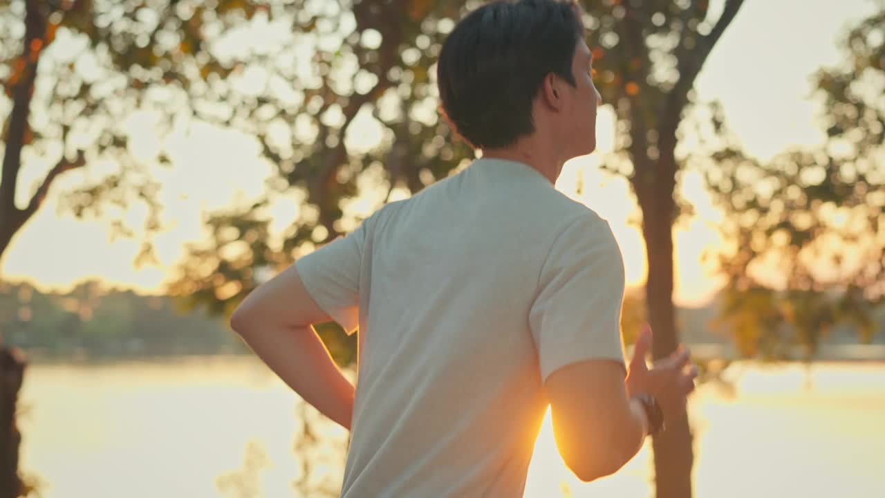 侧视图-有吸引力的亚洲男子慢跑在公园的树木背景与阳光在日落，健身男子跑步，训练跑步锻炼。视频素材