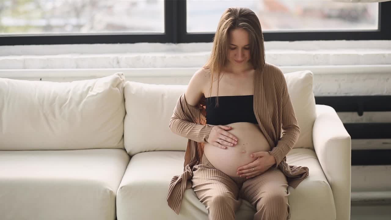 怀孕的快乐微笑的女人坐在沙发上说话，抚摸着她的肚子。妈妈要生孩子了。孕妇的肚子。怀孕。美丽的孕妇。母亲的概念。视频下载