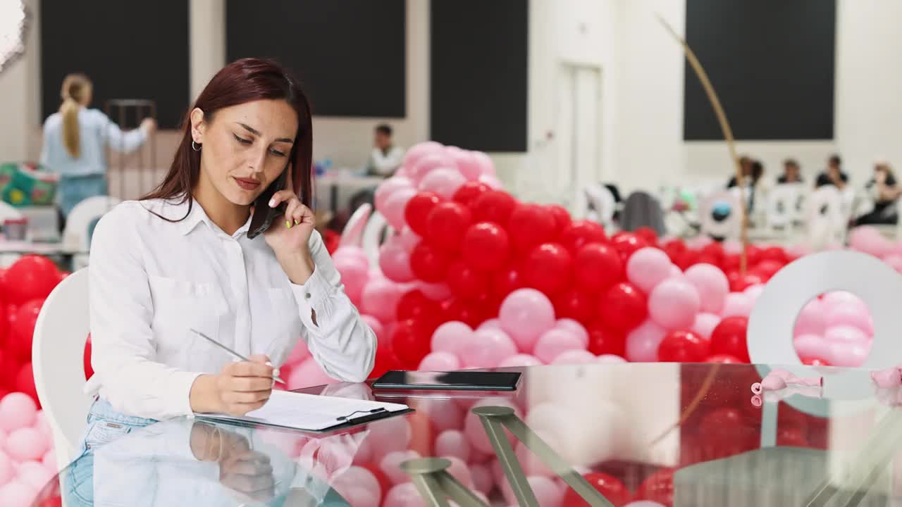 一位无忧无虑的女性活动组织者坐在餐厅里，与新客户通电话，写下客户想要的预订细节和装饰视频下载