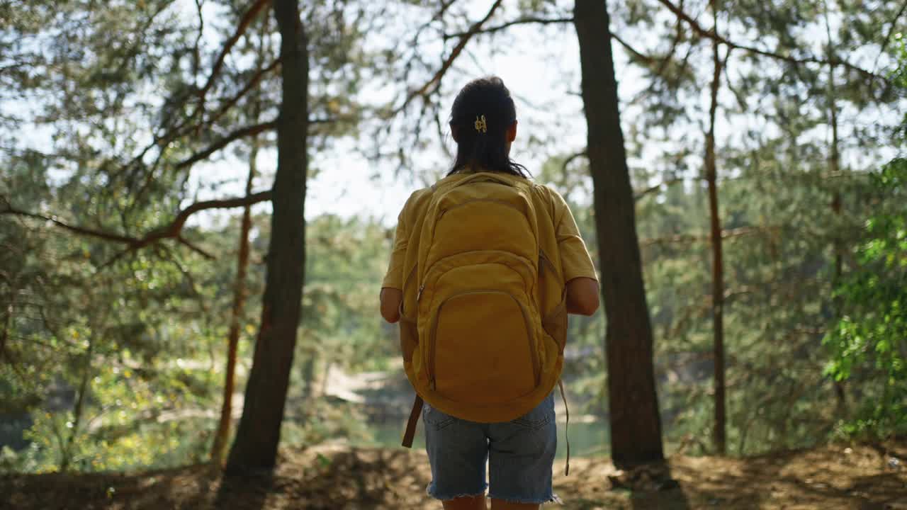 在一个阳光明媚的秋日里，女游客走在秋天森林里的小路上。在阳光明媚的早晨，背着黄色背包的女性徒步旅行者在森林里绕着山湖徒步旅行，冒险的户外生活方式视频下载