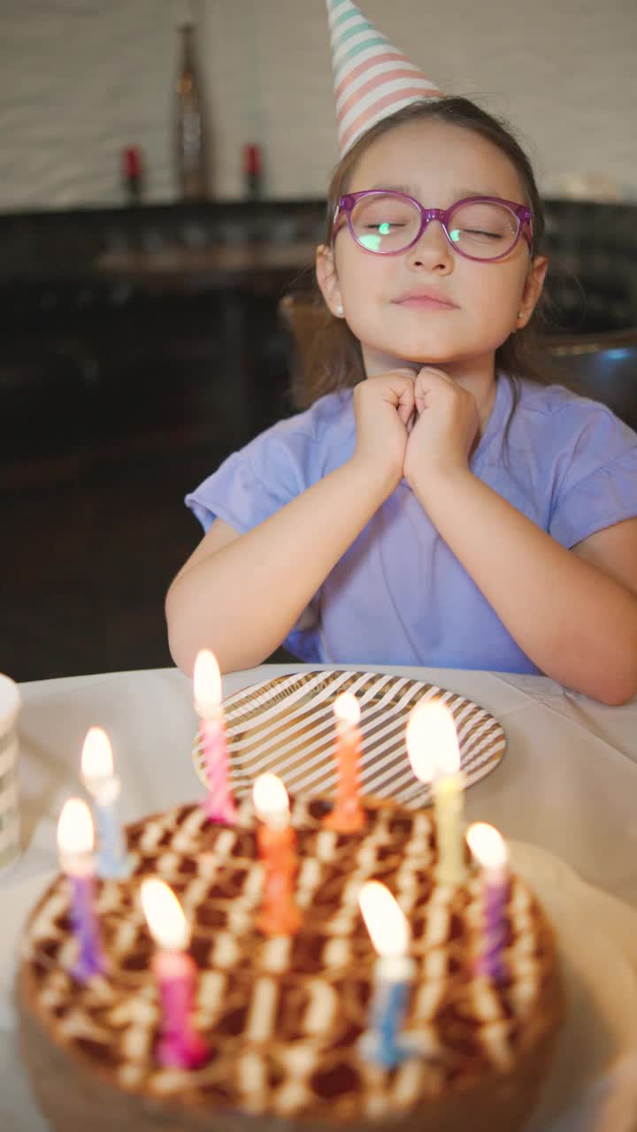 一个穿着紫色t恤，戴着彩色眼镜的小女孩正在为她的生日许愿，并吹灭了蛋糕上的彩色蜡烛视频素材