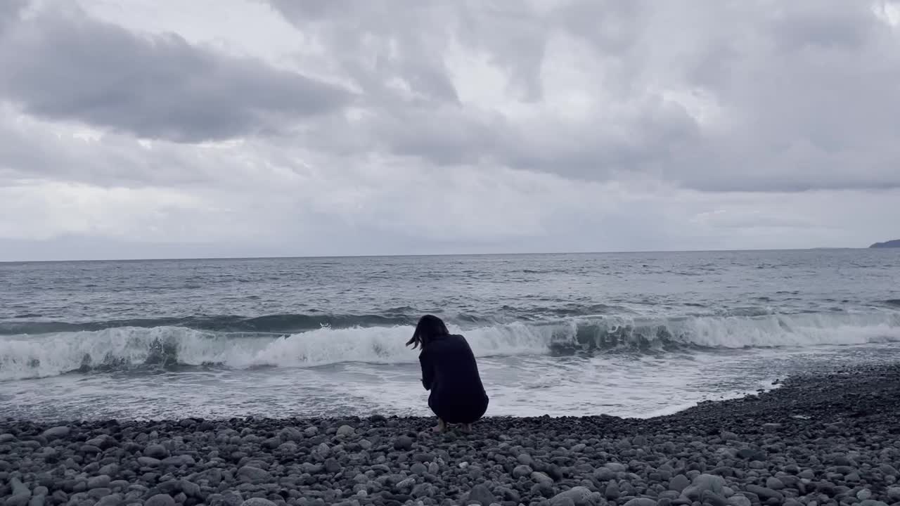 在阴天的海滩上，一个年轻女子闷闷不乐地蹲着视频下载