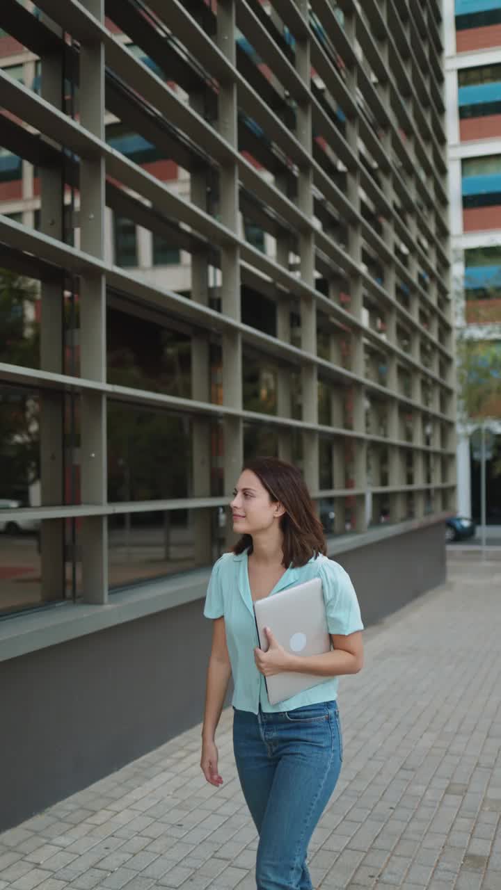 垂直视频，一个穿着休闲装的商务女性走在城市里，走进办公室，手里拿着笔记本电脑开始一天的工作视频素材