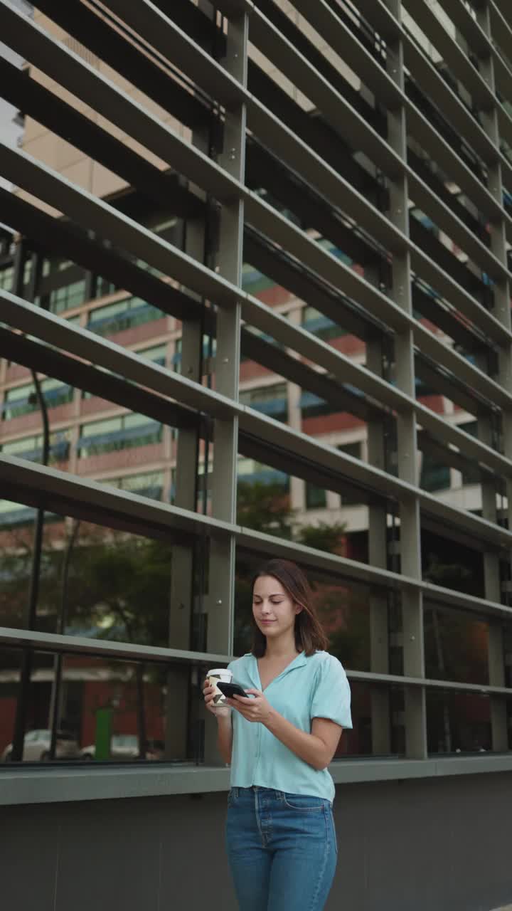 垂直视频，一位年轻的商务女性穿着休闲服，在办公室外工作休息时拿着咖啡，用手机视频素材