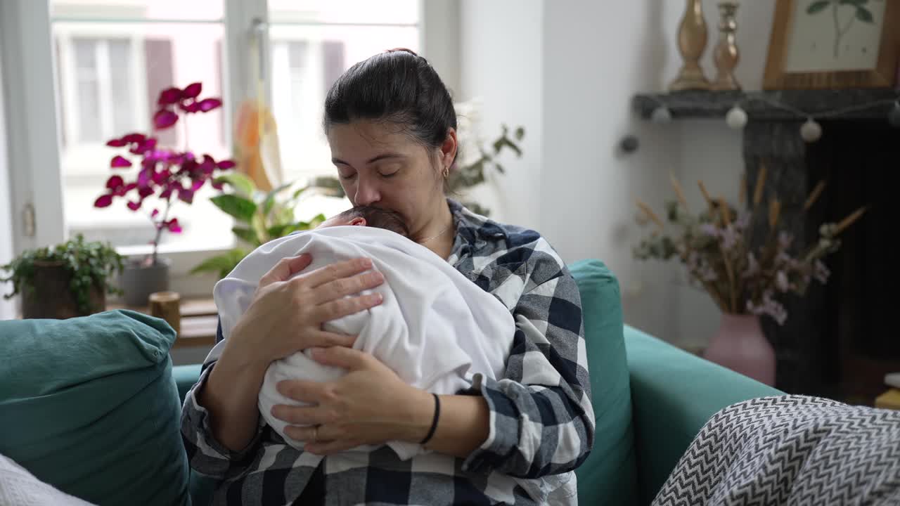 母亲抱着用毯子裹着的新生婴儿，坐在沙发上，看起来很关心，在一个舒适的家庭环境中，捕捉到一刻的关怀和母爱视频素材