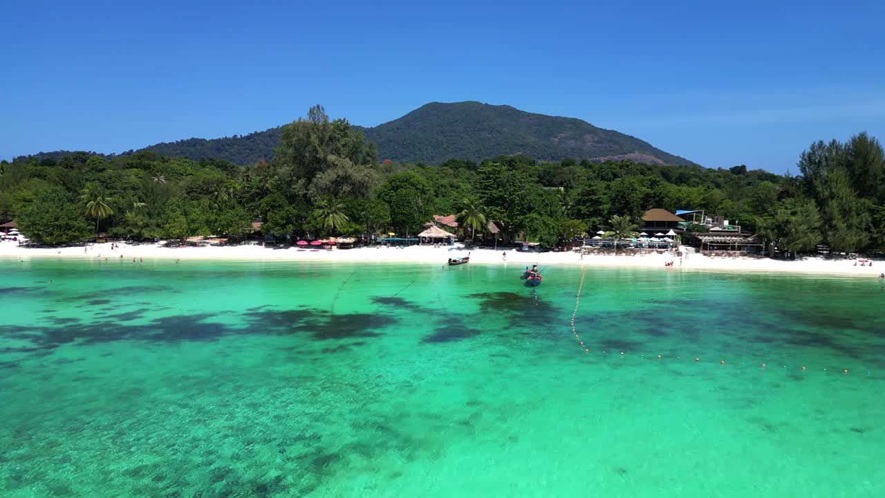 游客们在热带岛屿海滩上享受阳光明媚的一天，传统的泰国长尾船停泊在碧绿的海水中。不错的鸟瞰图飞行无人机全景概述无人机视频素材