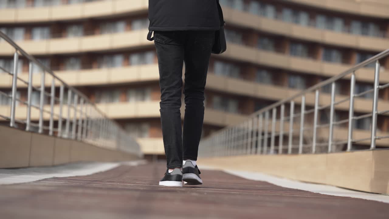 一个年轻人背着背包走在桥上。这个人穿着黑色的裤子和黑色的夹克。画面有一种孤独和沉思的情绪，慢动作视频下载