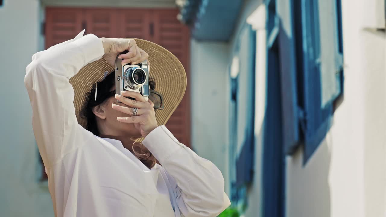 戴着帽子和眼镜的时尚模特在希腊雅典卫城下的阿纳菲奥蒂卡狭窄的街道上漫步，拿着复古相机拍照视频下载