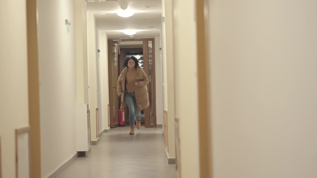 一头卷发的西班牙裔女子穿着冬季夹克和蓝色牛仔裤在大楼走廊里奔跑，慢镜头视频素材
