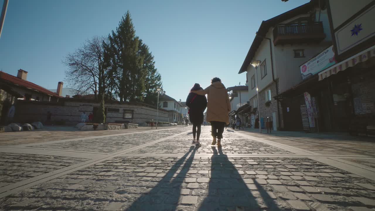保加利亚，班斯科——2024年2月25日:在保加利亚班斯科，两名女游客在阳光的照射下走在皮林街的人行道上，慢动作视频下载