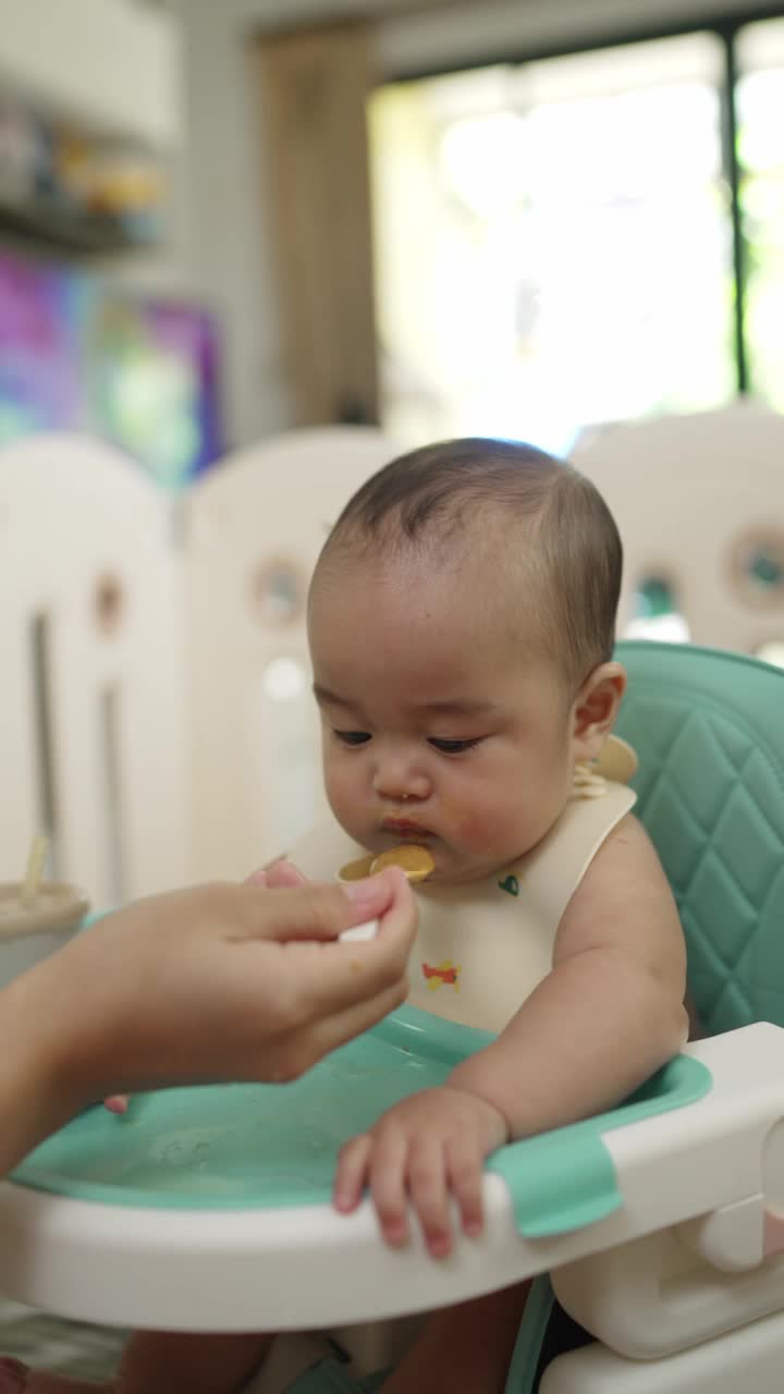 垂直视频。一个婴儿正在用杯子吃东西，而一个人在旁边看着。婴儿戴着围嘴，坐在一张高椅子上视频素材