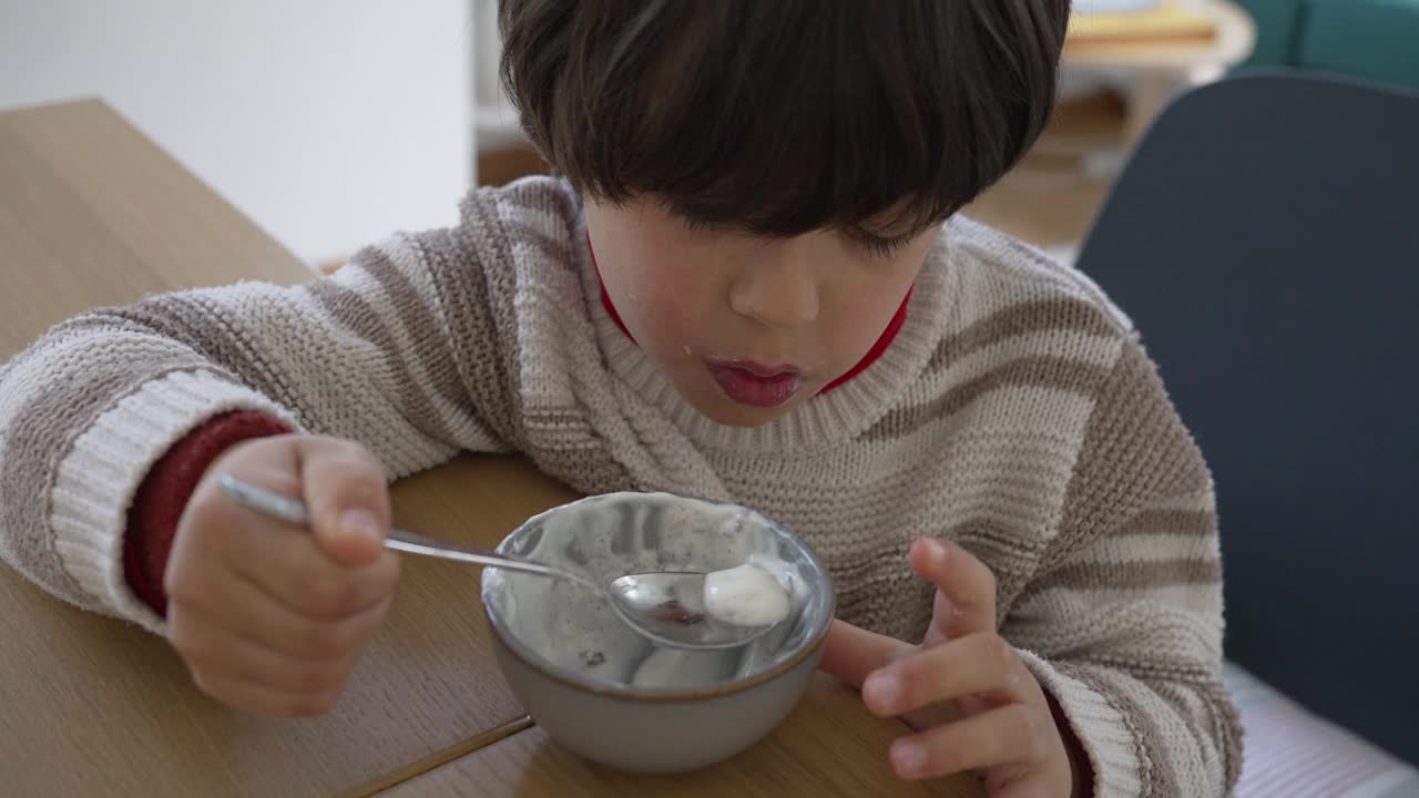 小男孩拿着勺子从碗里吃酸奶，全身心地投入到活动中，描绘了一个率真而自然的童年时刻在家里，气氛轻松宁静视频素材