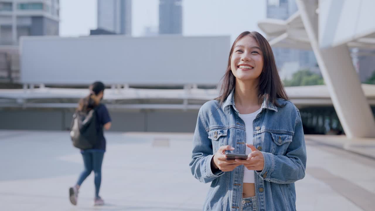 年轻的亚洲女子穿着牛仔裤，夹克，衣服，在晴朗的日子里走在城市的街道上，用智能手机发短信视频素材