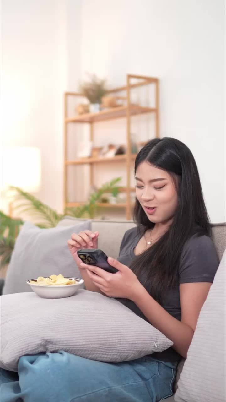 一位年轻的亚洲女子坐在沙发上，一边用智能手机一边吃着碗里的薯片。客厅里有一株植物和一盏灯作为背景视频素材