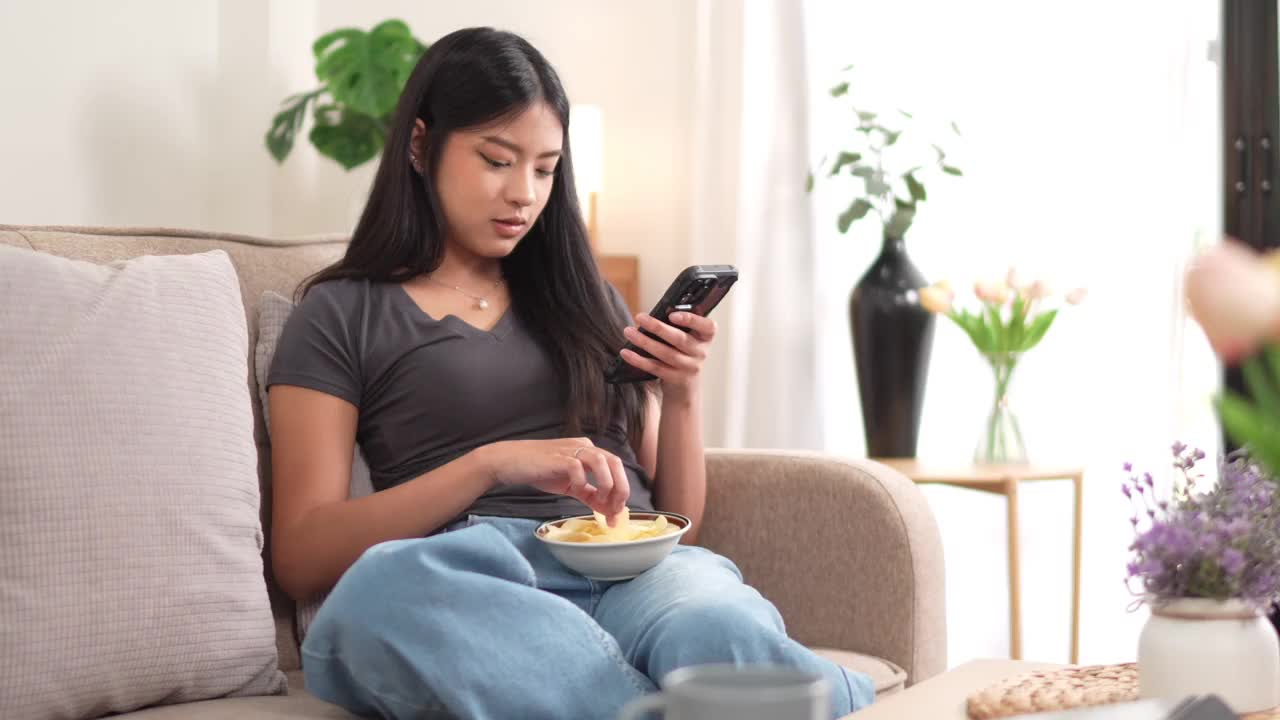 一位年轻的亚洲女子坐在沙发上，一边用智能手机一边吃着碗里的薯片。客厅里有一株植物和一盏灯作为背景视频素材