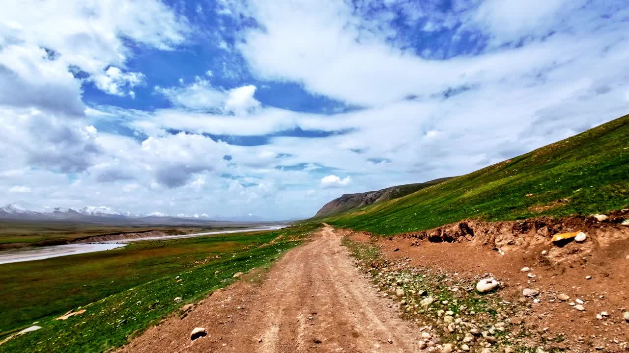 沿着绿油油的高河岸的大江山行驶在土山路上。背景是白雪皑皑的山脊。天气晴朗，天空多云视频素材