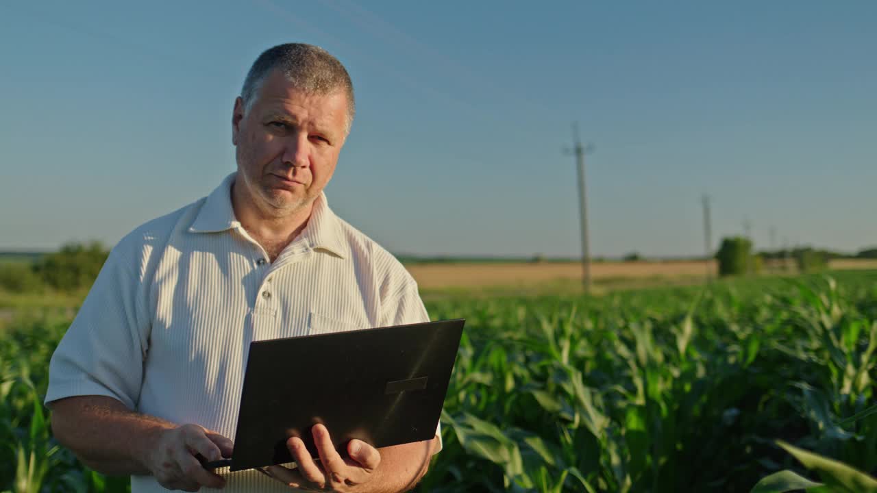 一个中年农民拿着笔记本电脑，站在风景如画的农村，玉米和小麦正在生长视频下载