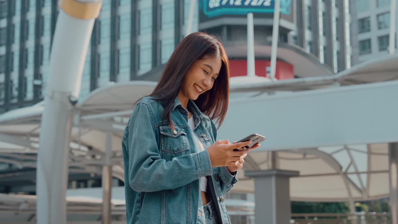 年轻的亚洲女子穿着牛仔裤，夹克，衣服，在晴朗的日子里走在城市的街道上，用智能手机发短信视频素材