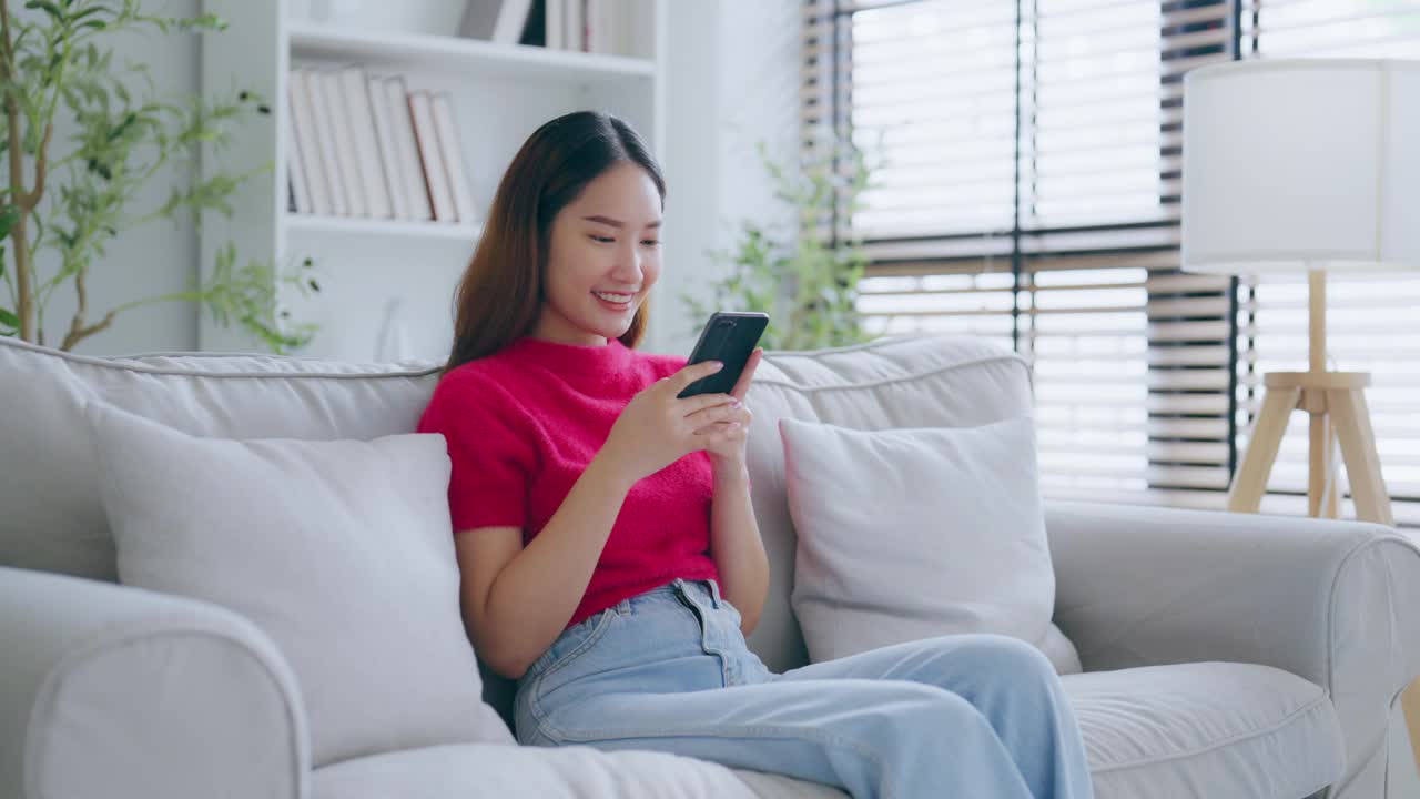 快乐的年轻亚洲女人放松在家里舒适的沙发上发短信在智能手机上，微笑的女孩使用手机，聊天在线消息，从家里网上购物视频素材