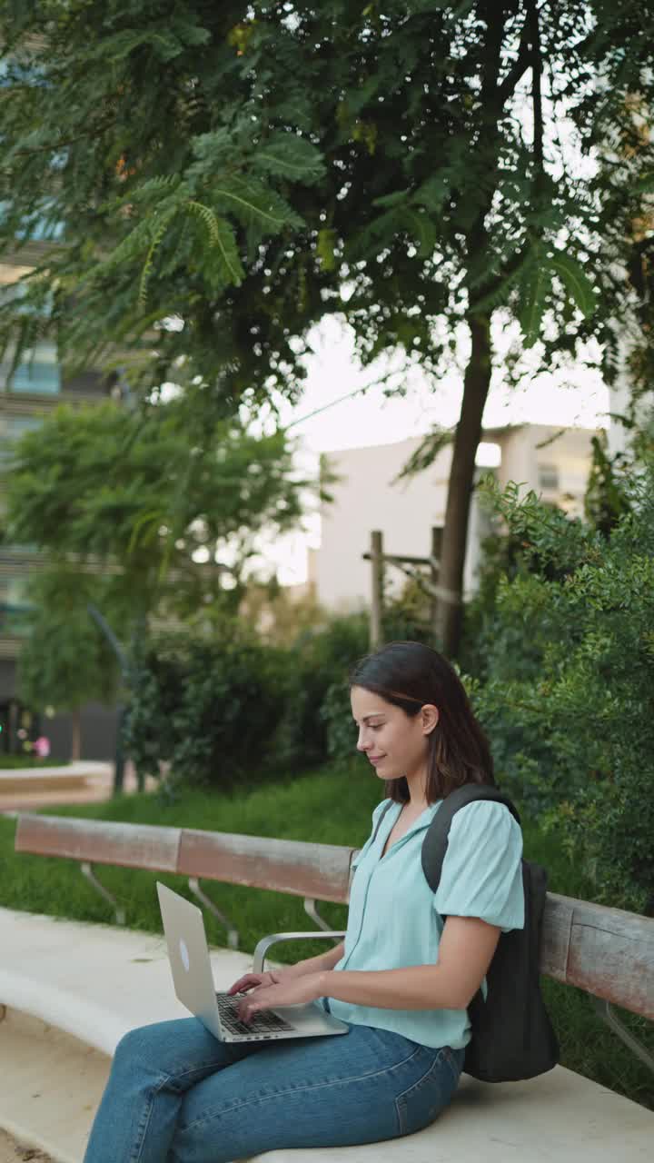 垂直视频显示，一名年轻的自由职业女性穿着休闲服，坐在城市公园的长椅上，对着镜头微笑着在笔记本电脑上工作。远程工作的概念视频素材