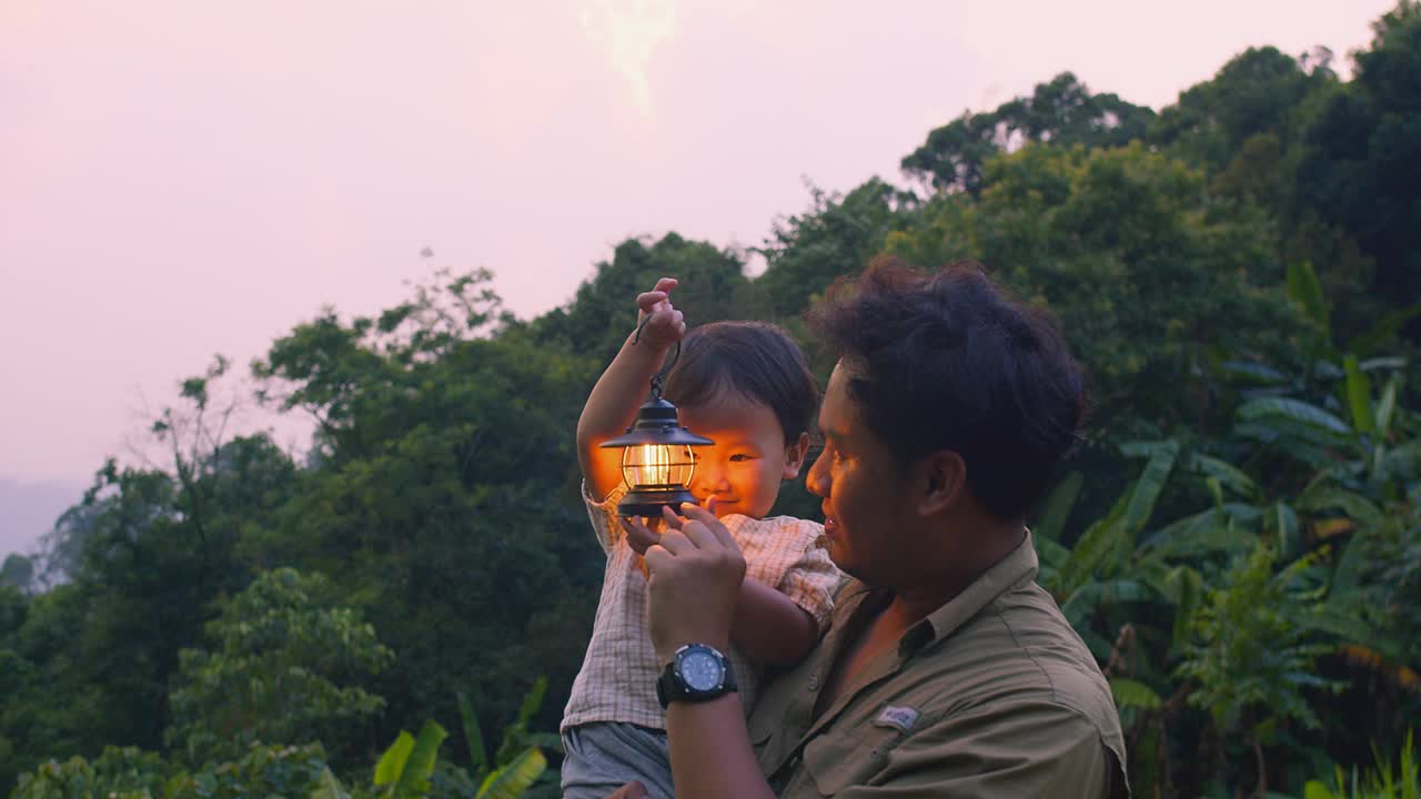 夕阳西下，父亲和孩子提着灯笼在郁郁葱葱的绿色森林中探险，营造出温暖而冒险的氛围。视频素材