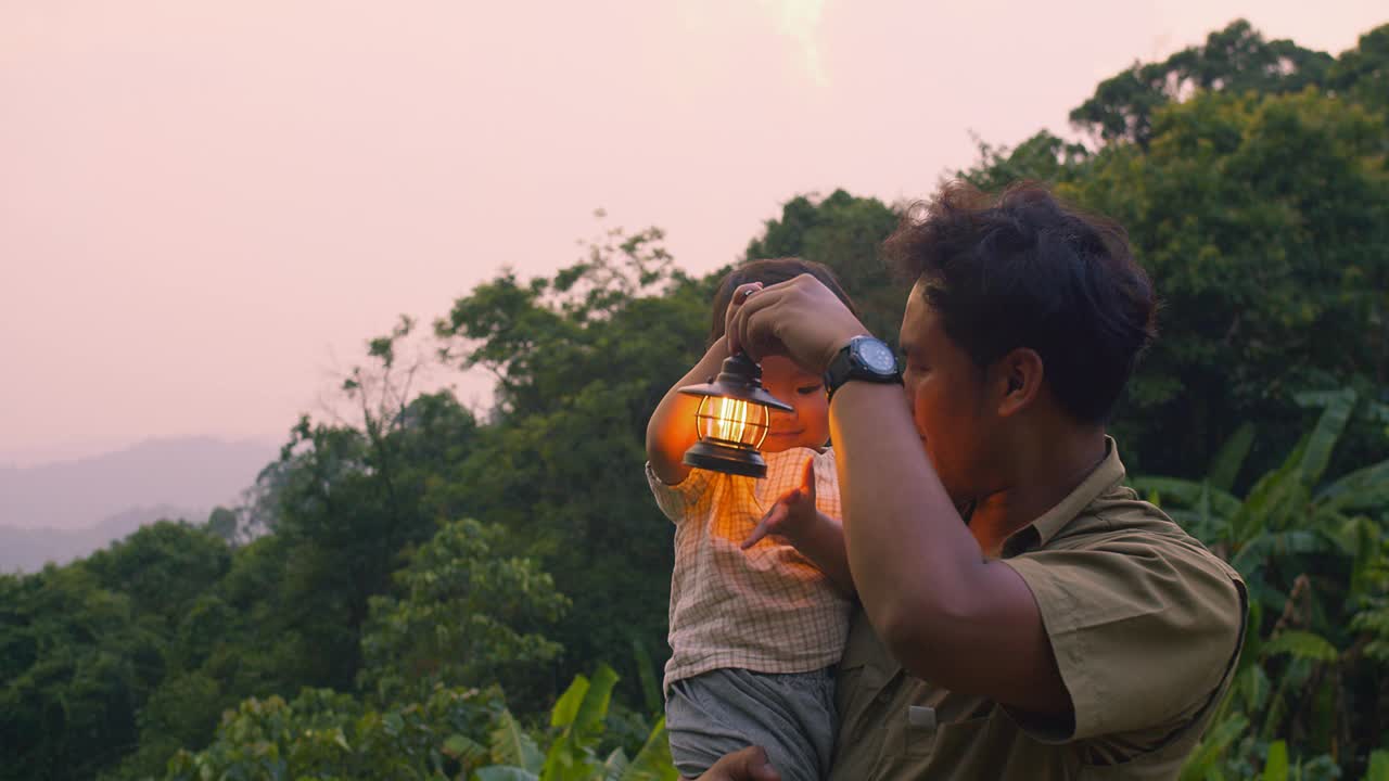 夕阳西下，父亲和孩子提着灯笼在郁郁葱葱的绿色森林中探险，营造出温暖而冒险的氛围。视频素材