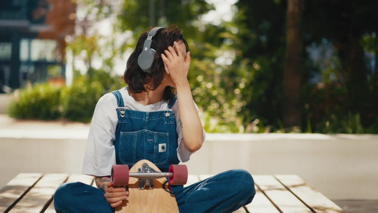 穿着牛仔工作服的女人坐在长凳上玩滑板，戴着耳机听音乐，享受夏日的阳光视频素材