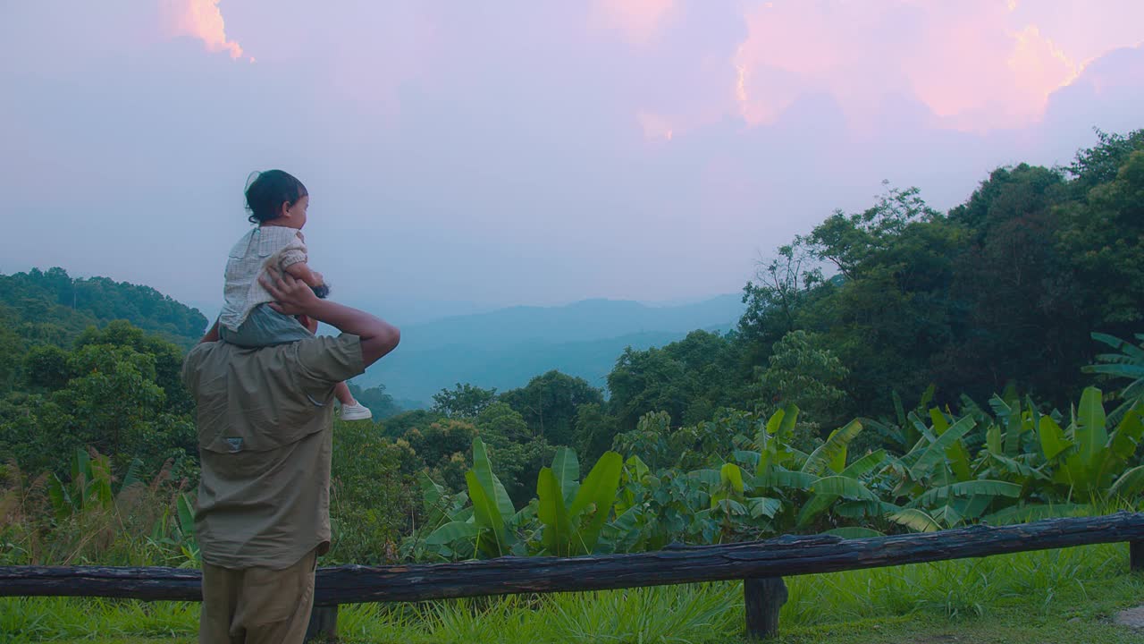 一位父亲带着他的孩子在他的肩膀上欣赏着美丽的山景，夕阳环绕着郁郁葱葱的绿色植物视频素材