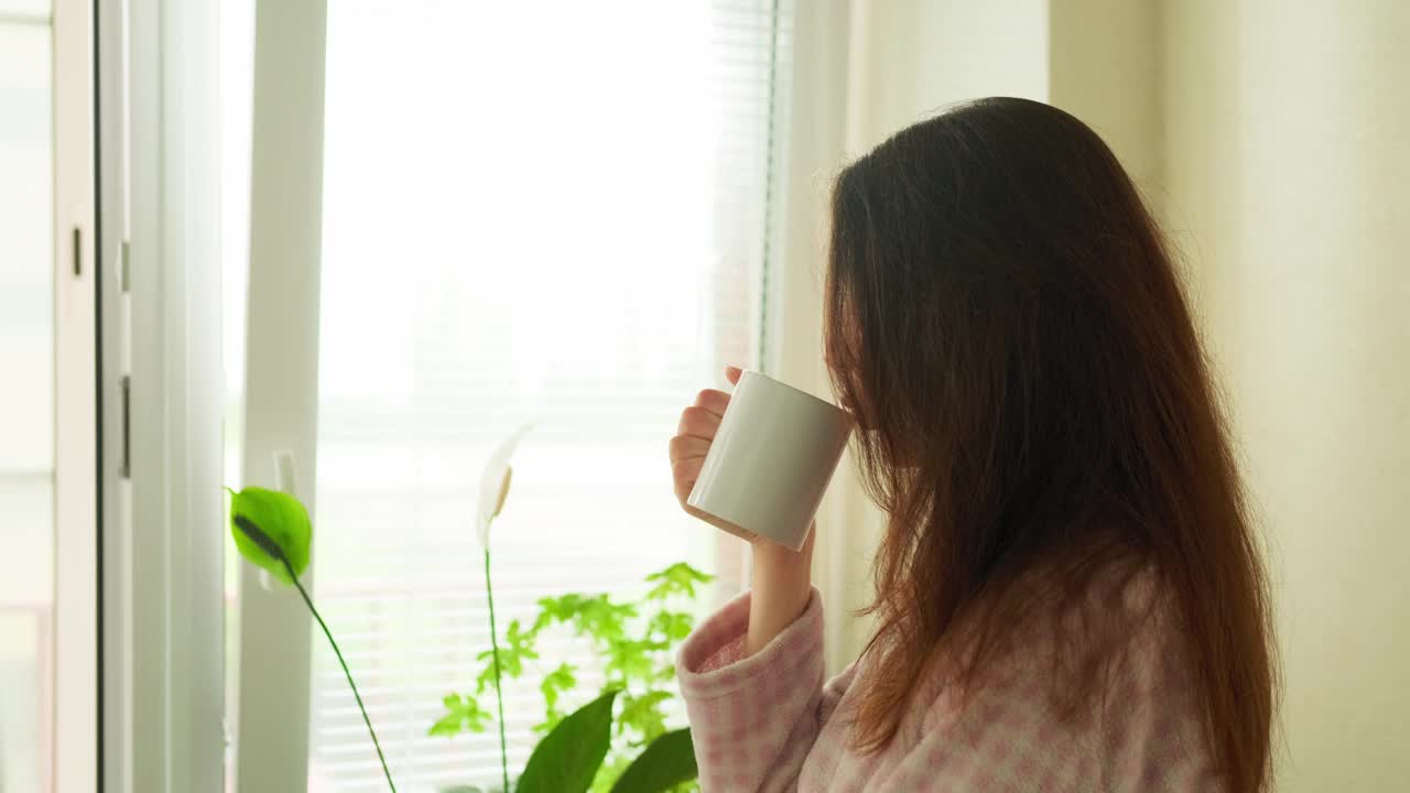 在一个阳光明媚的早晨，一个迷人的女人双手捧着杯子，在厨房里喝了一口咖啡，直直地望着窗外视频下载