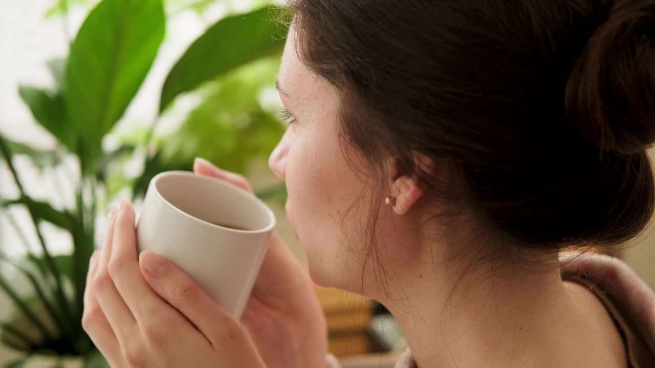 一个阳光明媚的早晨，一个迷人的黑发女人在厨房里手捧着杯子，喝了一口咖啡，眼睛直视窗外。后视图视频下载