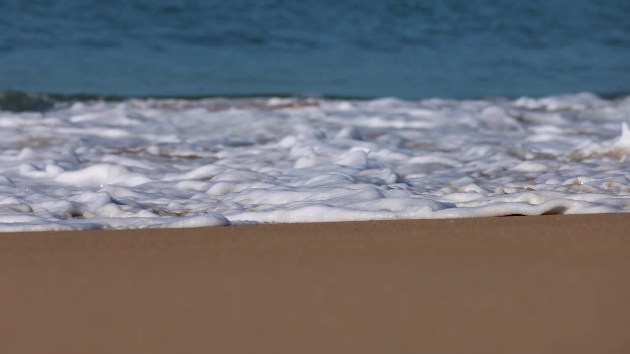 一个泡沫的波浪沿着沙滩海岸线滚动的特写镜头，从低角度拍摄慢动作。视频下载