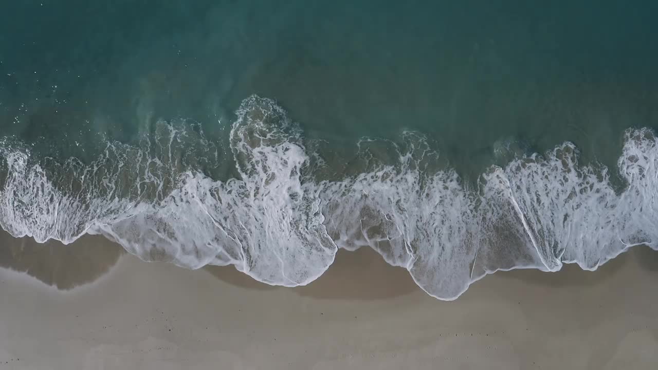 多个波浪沿着浅沙海岸线破碎，创造出抽象的移动纹理。当相机平行于海滩移动时从上面拍摄。视频素材