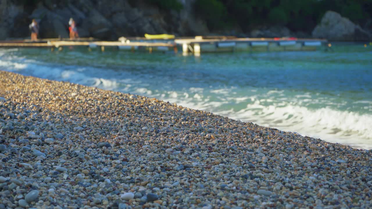 特写细节——海滩上的小鹅卵石或粗糙的沙子，背景是模糊的下午阳光照亮的平静的大海，有船的木制码头视频下载