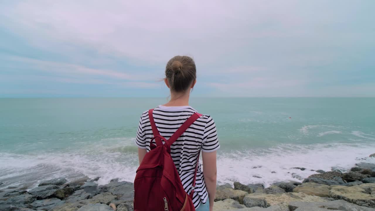 女人站在多岩石的海岸上，看着惊涛骇浪的背影视频素材