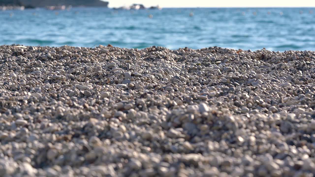 特写细节——海滩上的小鹅卵石或粗糙的沙子，背景是模糊的下午阳光照亮的平静的大海，相机慢慢地转动视频下载