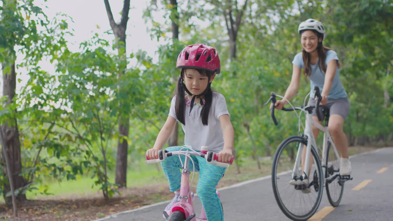 亚洲年轻的母亲和女儿在户外花园里骑自行车。快乐的家庭，漂亮的妈妈穿着运动服，心情愉快，放松，在公园里和可爱的小女孩一起锻炼身体。视频下载