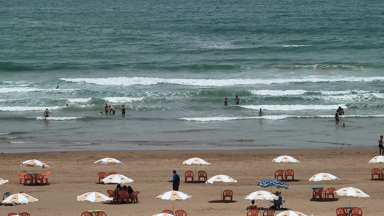 一个风景如画的海滩，沙滩上有单色的雨伞和椅子可供出租。人们沿着海岸线游泳和散步视频素材