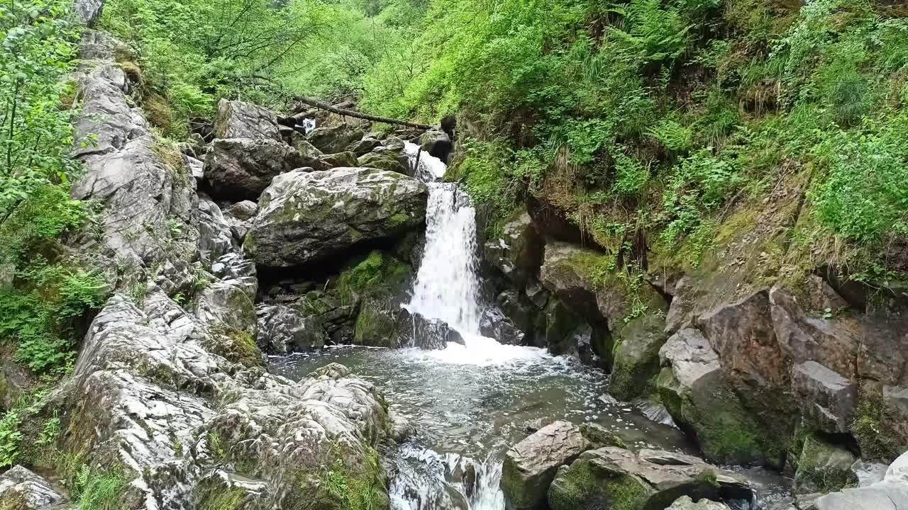 在夏日的森林里，一条小瀑布从山间的裂缝中流下，撞击着湍急的溪流中的石头。视频下载