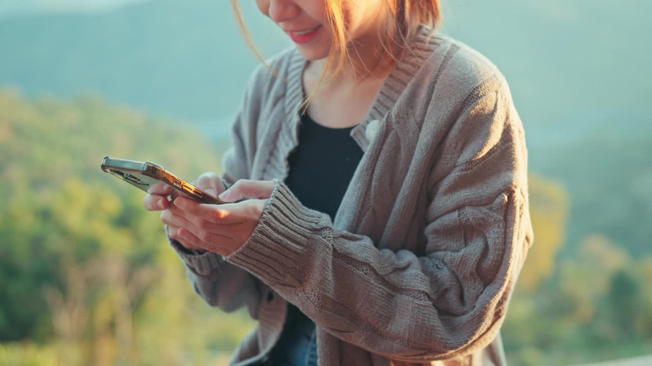 特写-快乐的亚洲妇女使用手机，滚动屏幕。女人早上坐在对着山的阳台上看社交媒体上的内容，购物。视频下载