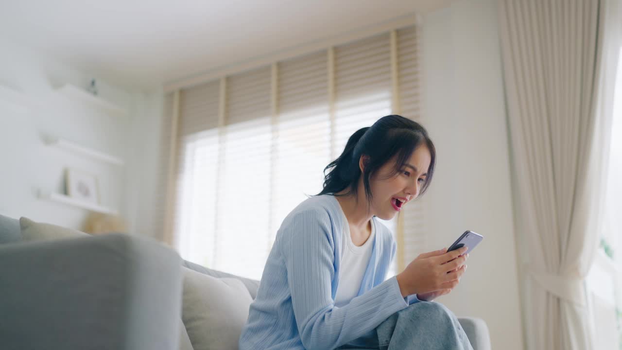 微笑的年轻漂亮的亚洲女人坐在舒适的沙发上，在客厅里使用有趣的手机应用程序。女人在家，网上购物，给朋友发短信，在社交媒体上发帖视频素材