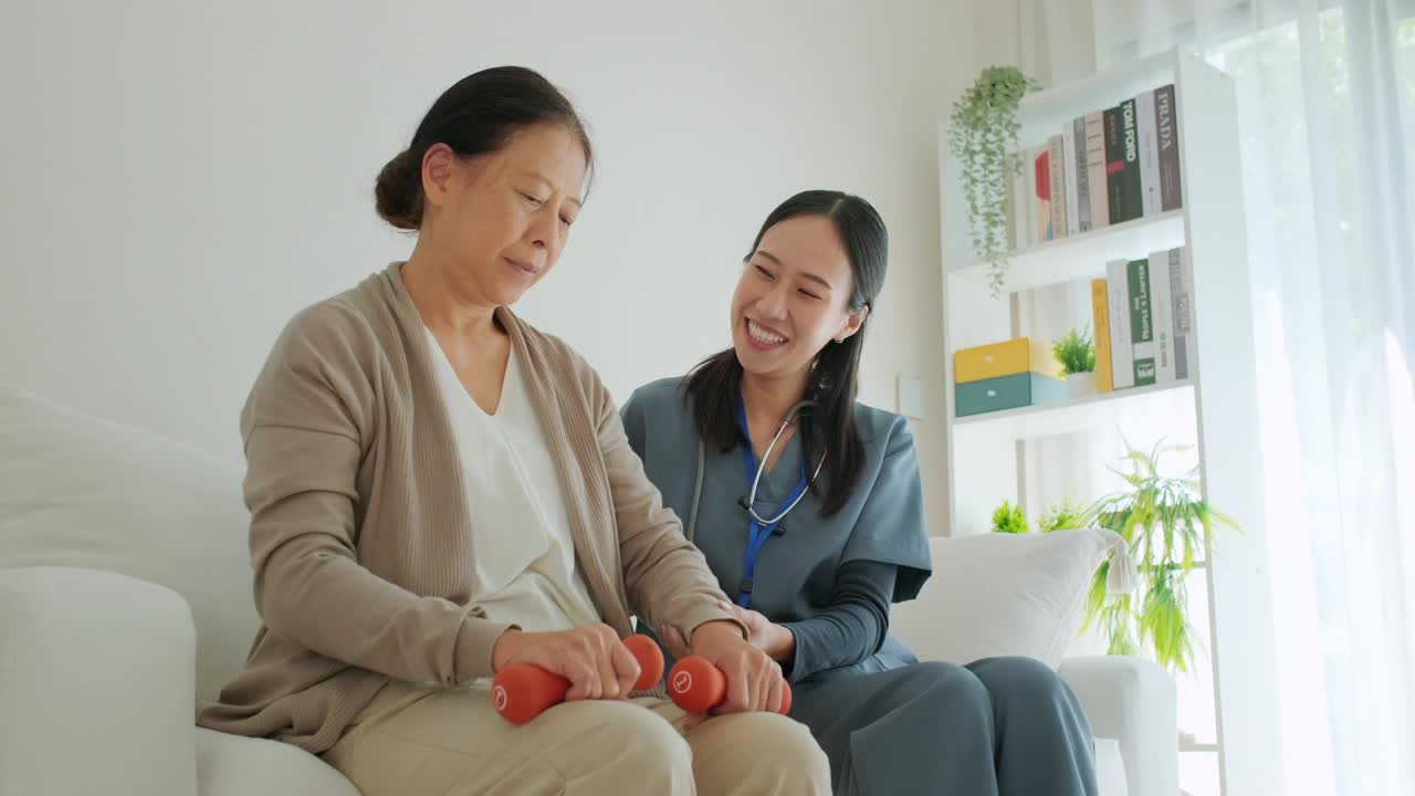 穿着蓝色工作服的亚洲护工在客厅帮助老妇人用哑铃进行轻度运动视频下载